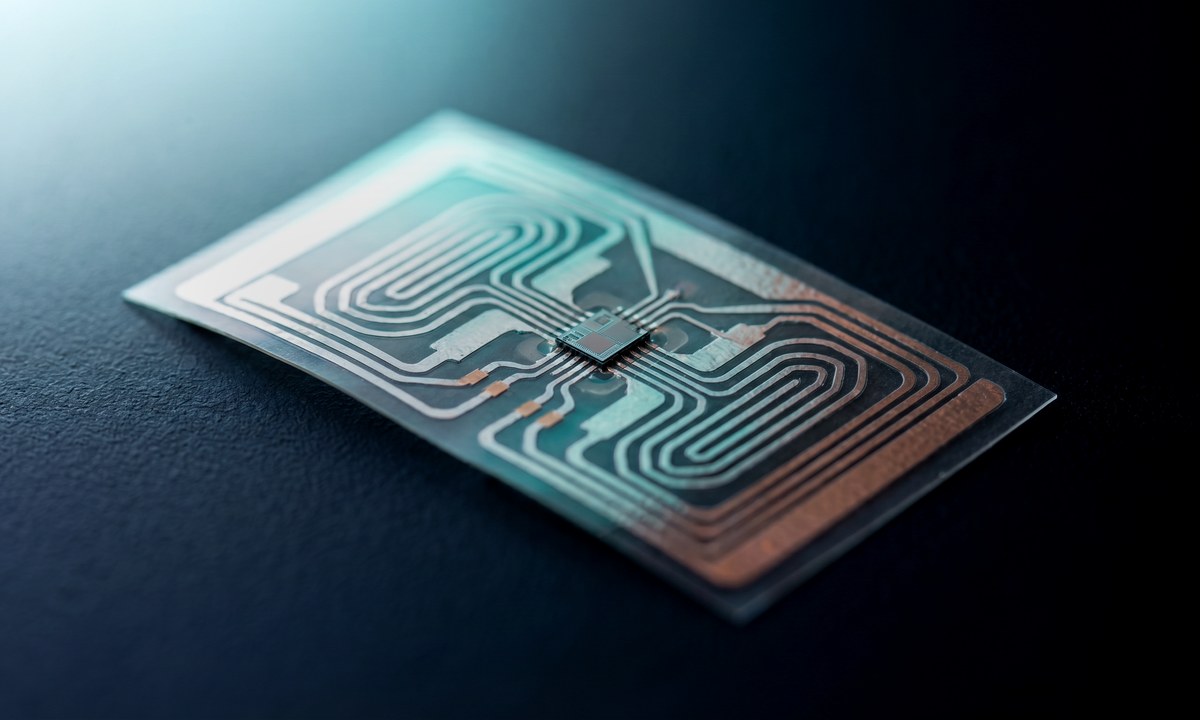 Exploded view of a printed flexible sensor inlay with a copper RFID antenna coil, silicon chip die at centre, and concentric energy harvesting waves